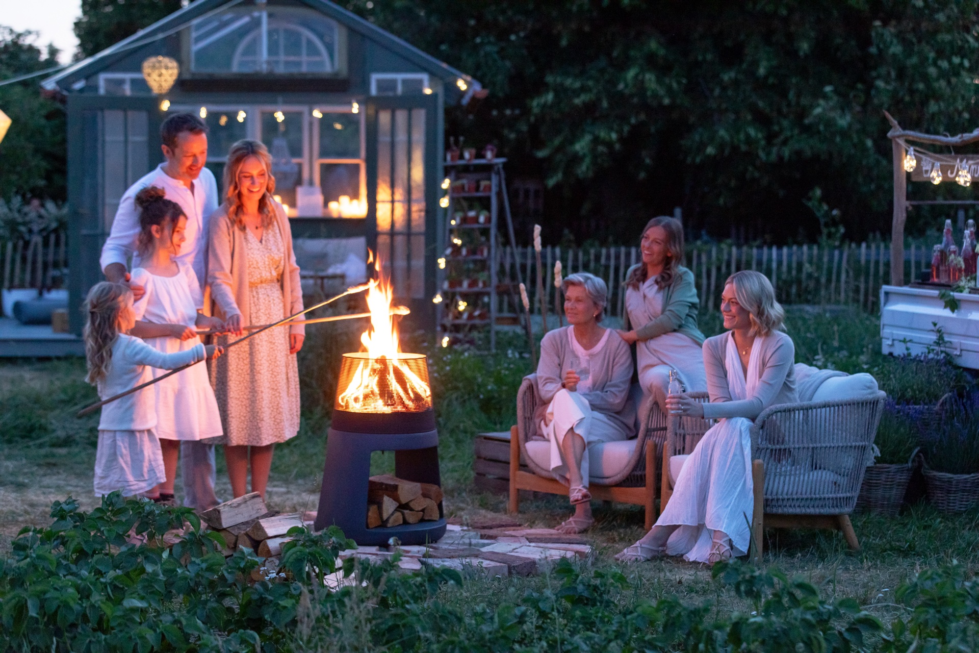 Kampagnen-Abend am Set: Menschen am Feuer im Garten, Lichterketten und Gewächshaus